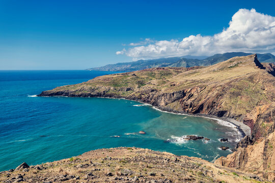 The photo depicts the picturesque landscape of Madeira PR8 Vereda da Ponta de São Lourenço: light blue sea, rocky coastline, and green hills under a blue sky.