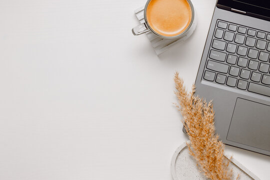 Minimal home office background with laptop, cup of coffee, tray and pampas with copy space on white wooden table, top view. Laptop workplace keyboard background, aesthetic style