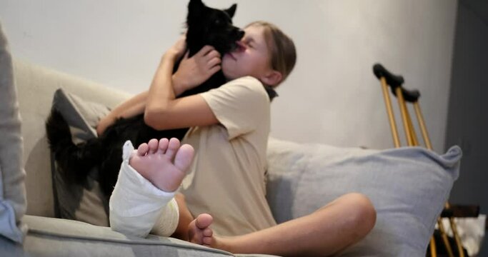 A teenage girl with a cast on her leg sits in the living room on the sofa and plays with a small black dog. A teenager has a broken leg with crutches in the background. Recovery after injury.