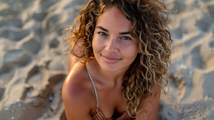 Woman with curly hair smiling and looking at camera while resting on sandy beach 
