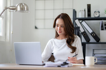 Young woman concentrating on her laptop in a well-lit home office setup with an open notebook, Thinking work concept.
