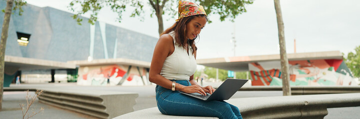 Cute tanned woman with long brown hair in white top and yellow bandana using laptop while sitting on bench. Pretty girl student preparing for classes in outside, Panorama