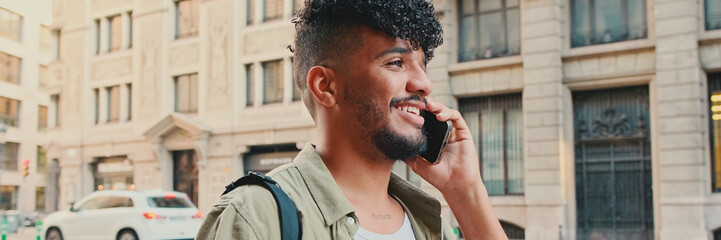 Young happy man with beard dressed in an olive-colored shirt is talking on cellphone standing on the old city background, Panorama