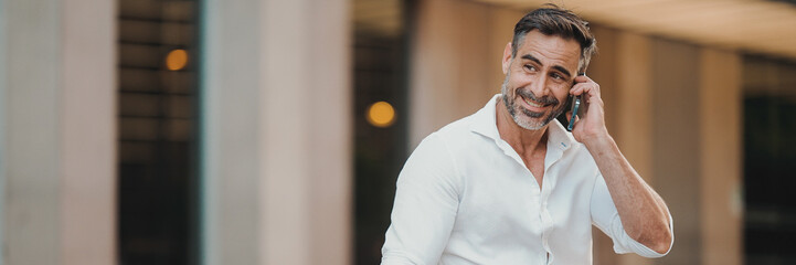 Mature businessman with neat beard wearing white shirt uses mobile phone sits on bench in the...