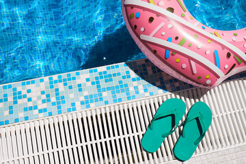 Pink inflatable ring and green rubber flip-flops by blue outdoor pool water. Poolside relaxation.