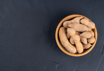 Wooden bowl filled with tamarind fruits on empty dark background, top view
