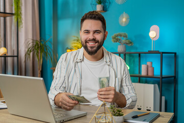 Happy business man counting money cash and using laptop in home office. Successful freelancer guy...