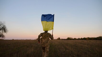 Soldier of ukrainian army running with raised blue-yellow banner on field at dusk. Young male military in uniform jogging with flag of Ukraine at meadow. Victory against russian aggression concept - 766465133