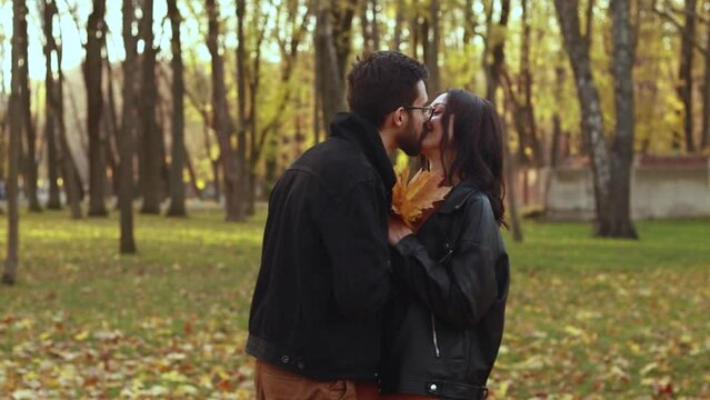 Young loving couple walking outside in the city park in sunny weather, collect orange yellow red maple leaves, hugging smiling kissing laughing spending time together. Autumn, fall season 
