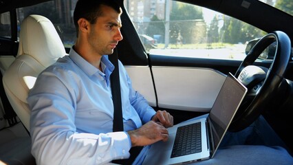 Handsome entrepreneur working on notebook while riding an autonomous self driving electric car at urban road. Male businessman typing text on laptop during riding on electrical vehicle with autopilot - 766467387