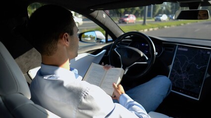 Male businessperson reading book during riding on electrical vehicle with autopilot at urban road. Successful businessman improving his knowledge while riding an autonomous self driving electric car - 766467968