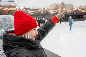 A woman in a red hat waves hello to friends at a skating rink in winter. 