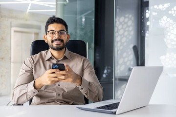 Confident Indian businessman in casual attire, using smartphone and laptop, sits in a modern office...