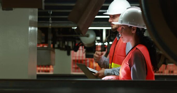 Male and female rail engineers, wearing orange safety vests and helmets, inspecting the underside of an electric train system in an industrial and engineering setting.