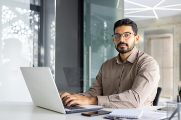 Professional Indian businessman in a modern office working intently on a laptop, portraying...