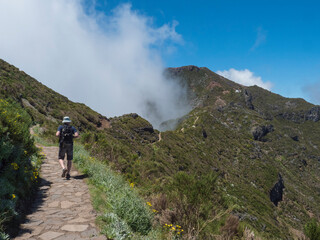 Man hiker in hat walking at paved footpath, hiking trail PR1.2 from Achada do Teixeira to Pico Ruivo mountain, the highest peak in the Madeira, Portugal. - 766493976