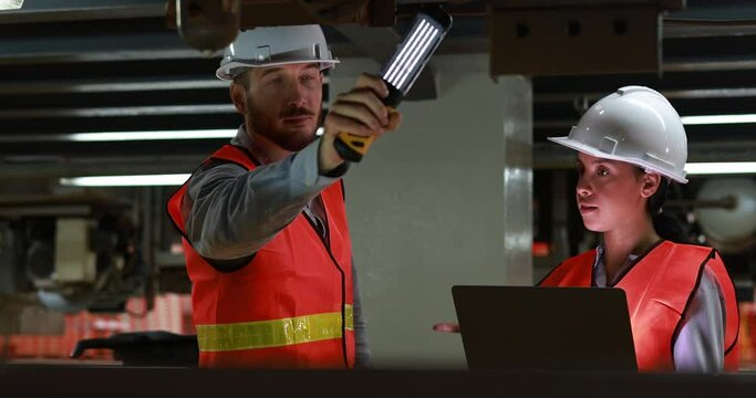 Male and female rail engineers, wearing orange safety vests and helmets, inspecting the underside of an electric train system in an industrial and engineering setting.
