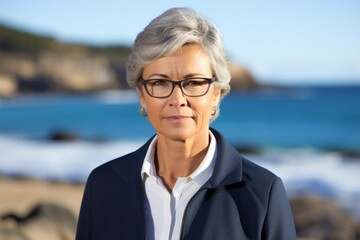 portrait of a middle aged woman with short gray hair wearing glasses and a blue jacket standing on a beach - Powered by Adobe