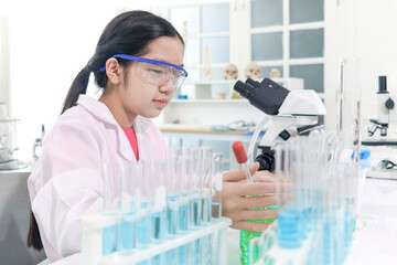 Cute young scientist schoolgirl wearing lab coat and safety glasses, doing science experiments. Student girl child using lab equipment to study chemistry in laboratory. Kid learning science education.