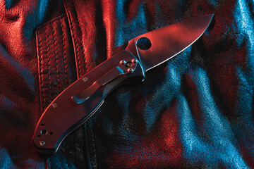 A folding knife lies on a leather jacket, multi-colored lighting.