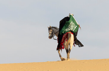 Saudi Man with his white stallion in a desert, carrying a flag of Saudi Arabia