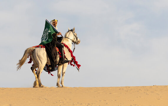 Saudi Man with his white stallion in a desert, carrying a flag of Saudi Arabia