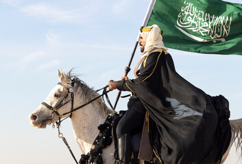 Man in traditional Saudi Arabian outfit and carrying a flag of KSA on a horse in a desert