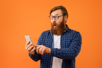 man with red hair excitedly browses on phone, orange background