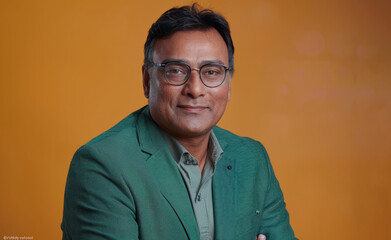 Fototapeta premium A man in a green and glasses is smiling for the camera. He looks happy and confident. Handsome happy Indian man in green business suit on bright orange colour background, colourful studio portrait