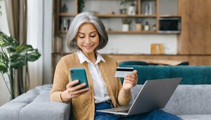 Serene middle-aged grey-haired Asian woman shopping online sitting with credit card and mobile phone at home, multiracial senior 40s female making order online, booking, transfering money