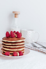 A stack of pancakes is decorated with raspberries and crushed almonds, in the background there is a bottle of milk and a white cup, a striped towel with black cutlery. on a white background