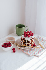 a stack of pancakes with vanilla sauce, decorated with raspberries and crushed almonds, next to a green cup of coffee and bowls with raspberries and sauce, a striped towel with gold cutlery. on a whit