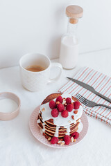 A stack of pancakes with vanilla sauce is decorated with raspberries and crushed almonds, in the background there is a bottle of milk and a white cup, a striped towel with black cutlery. on a white ba