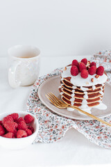 a stack of pancakes with vanilla sauce, decorated with raspberries and crushed almonds, behind a white cup with coffee and a bowl with raspberries. a colorful towel with gold cutlery. on a white backg