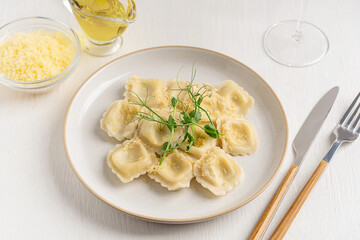 Traditional italian Ravioli stuffed pasta filled with mozzarella cheese decorated with green fresh pea microgreens served on plate with tableware, olive oil and glass of wine on white wooden table