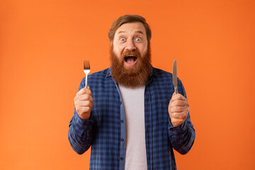 Hungry bearded man holds knife and fork expressing excitement, studio