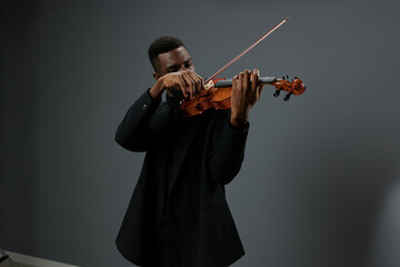 Talented young African American man in black suit performing classical music on violin against grey...