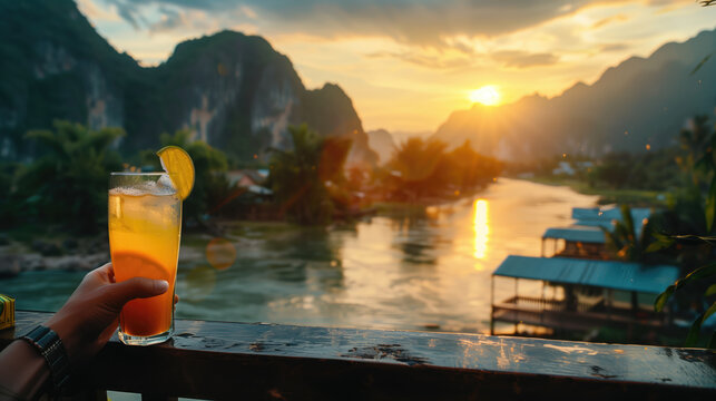 Hand holding a refreshing drink against a scenic sunset backdrop over a tranquil river surrounded by mountains.