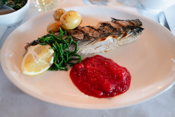 Cooked grilled mackerel fish on a plate with a raspberry and apple sauce. There is also some samphire a chuck of lemon and new potatoes.