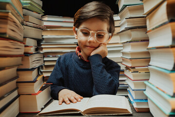 Cute little pupil boy in glasses reading interesting book in library between stacks of books literature. Education concept, prep or elementary school