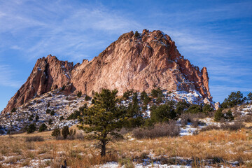 Garden of the Gods in Colorado Springs.  Red rock and blue sky. Tall red stone formations with light snow.  Natural red rock formations in Colorado winter.  Trees and desert scrub with snow foreground