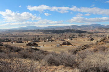 A winter view of countryside west of Devil's Backbone in Larimer County with blue sky and puffy white clouds.  Brown grasses and scrub bushes with rocky outcroppings and clouds.