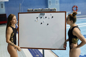 Two young female water polo athletes standing by blank whiteboard with magnets while discussing...