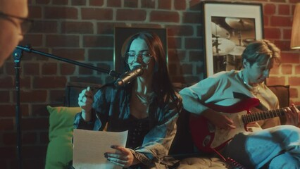 Medium footage of Caucasian female vocalist of musical indie band singing into mic to guitar and reading lyrics while recording song with friends at retro studio in evening