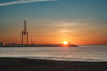 Puerto de Málaga, sol saliendo desde el horizonte sobre el faro del puerto, luz dorada, primeros...