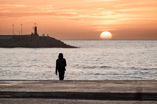 Playa de Málaga, hombre mirando el amanecer, primeros rallos de sol, en el horizonte el sol saliendo desde el mar, vista del faro del puerto de Málaga