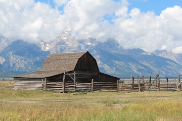 Old farm property in Wyoming.  Blue sky and green grass.  Clouds in sky.  Barn outbuilding in Wyoming countryside with Tetons in the background.  Mormon Row outbuildings. Tetons in August. 