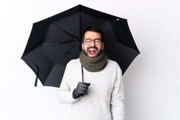Caucasian handsome man with beard holding an umbrella over isolated white wall shouting to the...