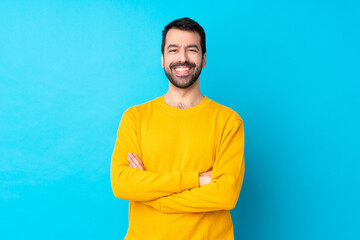 Young caucasian man over isolated blue background keeping the arms crossed in frontal position - obrazy, fototapety, plakaty