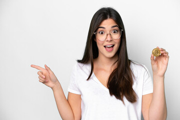 Young Brazilian woman holding a Bitcoin isolated on white background surprised and pointing finger...